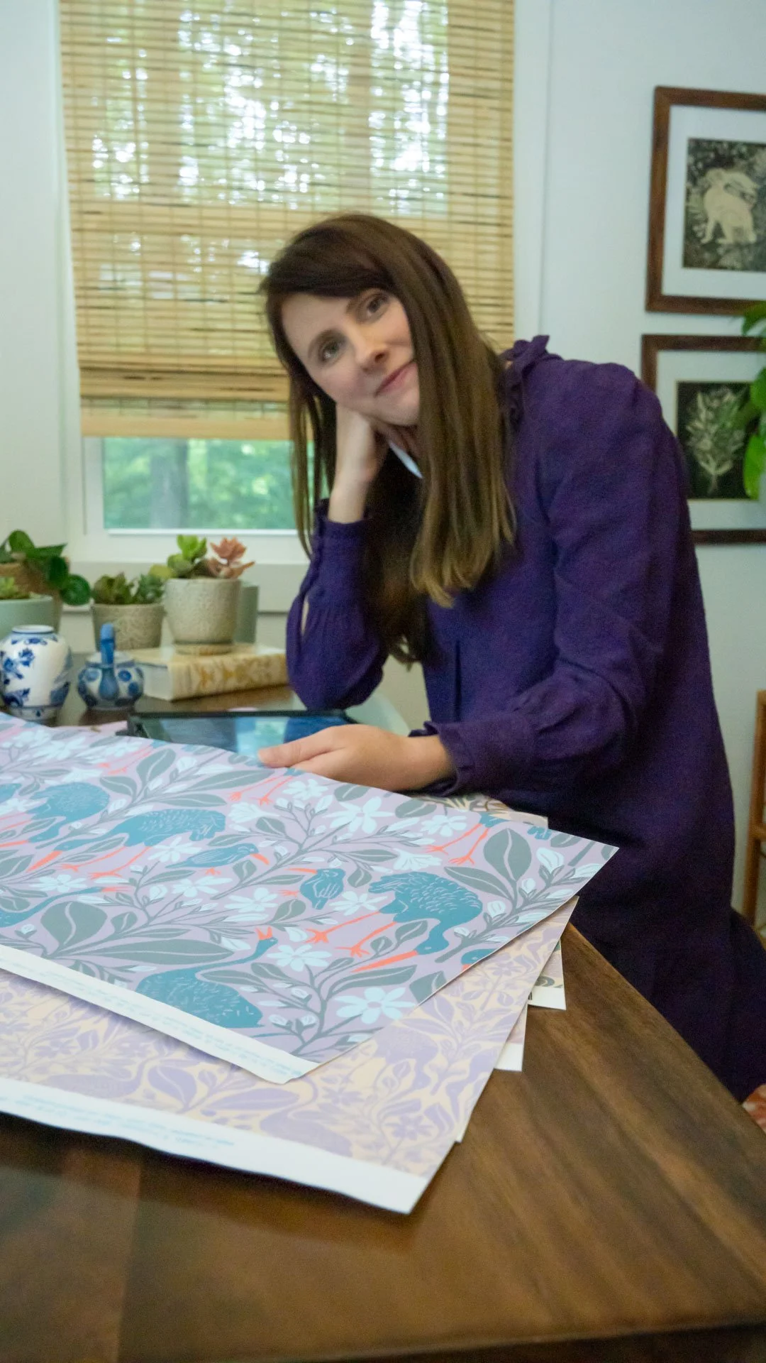 A woman with long brown hair, wearing a purple dress, is sitting at a wooden table with plants and decorative items in the background. She rests her head on her left hand and is looking at a tablet.