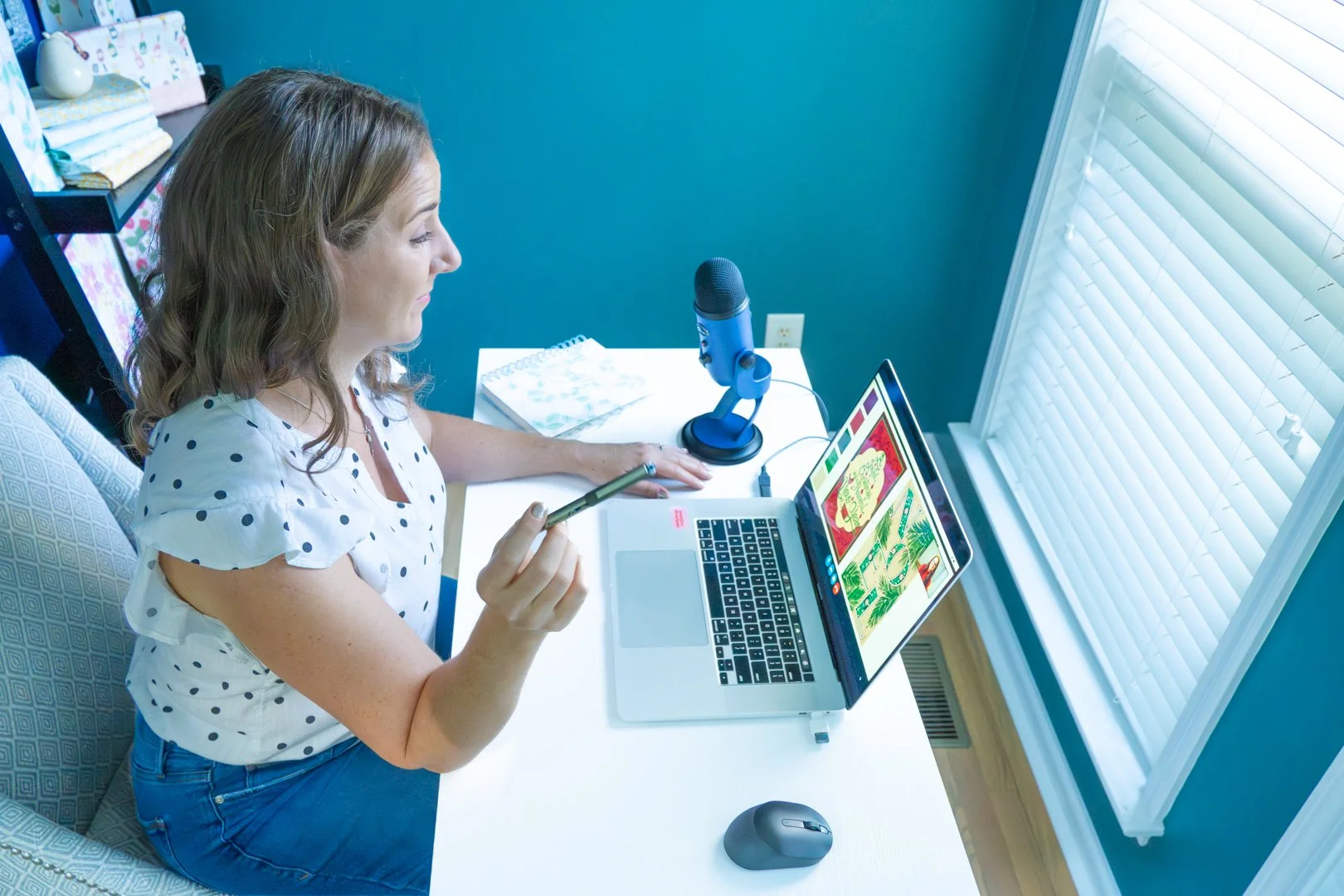Woman sitting at a desk using a laptop with colorful images on the screen, holding a stylus pen, in a room with teal walls and a window with blinds.