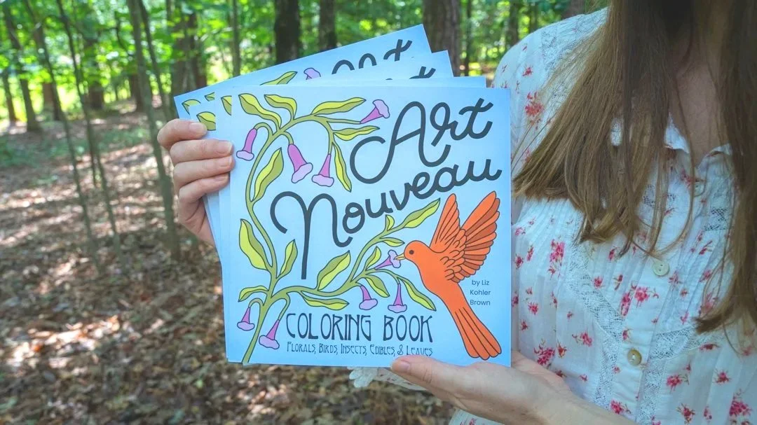 A person holding three copies of a coloring book titled 'Art Nouveau Coloring Book' with floral and bird illustrations.