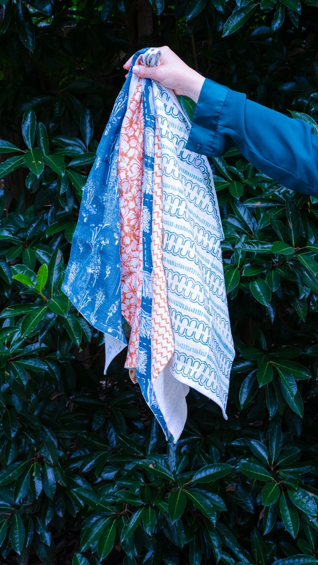 A hand holds several colorful fabric pieces with different patterns, hanging against a background of dark green leaves.