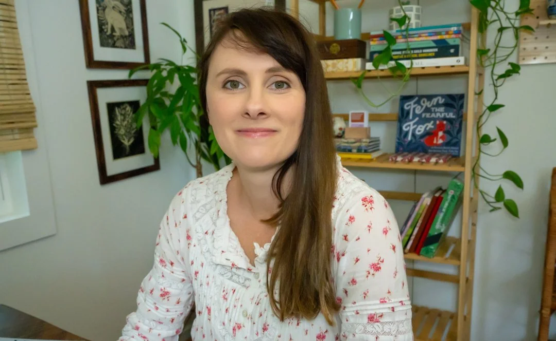 A woman with long brown hair and blue eyes smiling at the camera in a room with bookshelves, plants, and framed pictures.