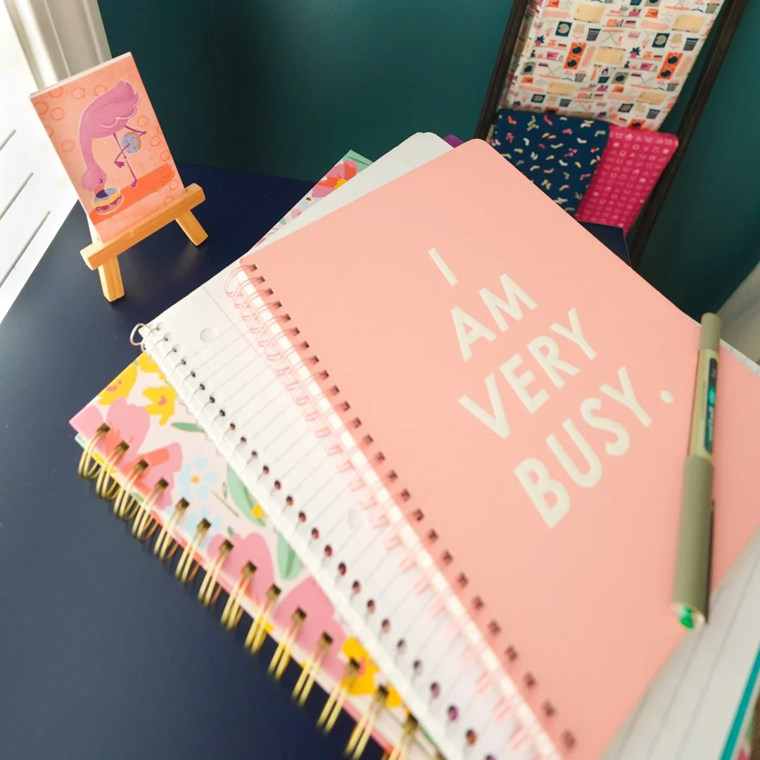 A desk with colorful notebooks, a pink notebook with the words 'I AM VERY BUSY,' a pen, and a small easel holding a cute illustration of a girl with pink hair drinking tea.
