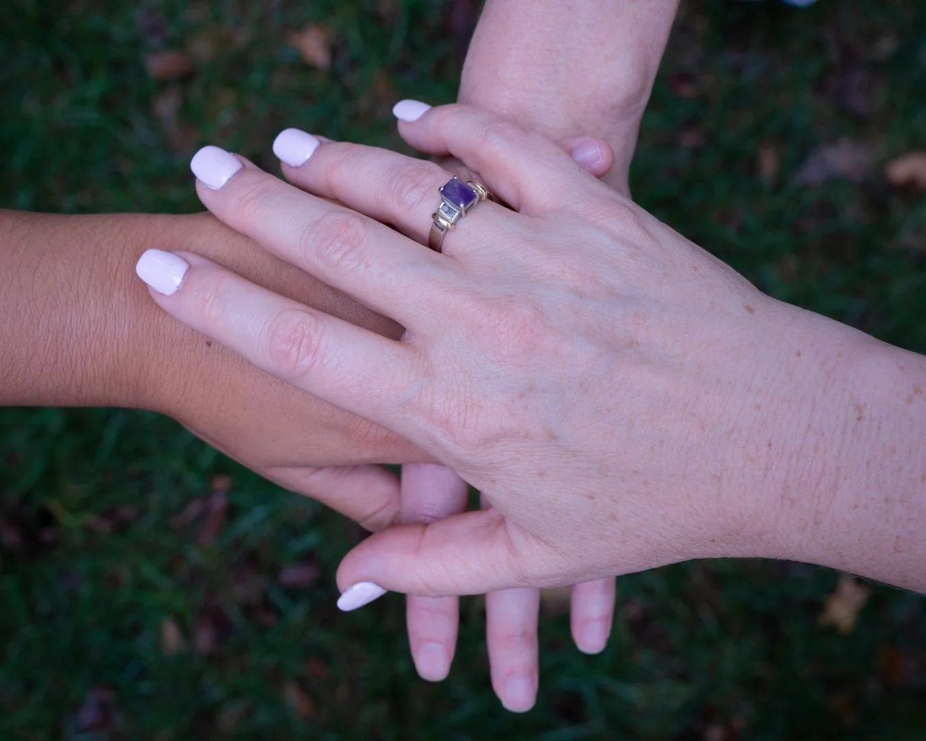 Two hands, one with a gold ring with a purple gemstone, touching each other's wrists, outdoors on grass.