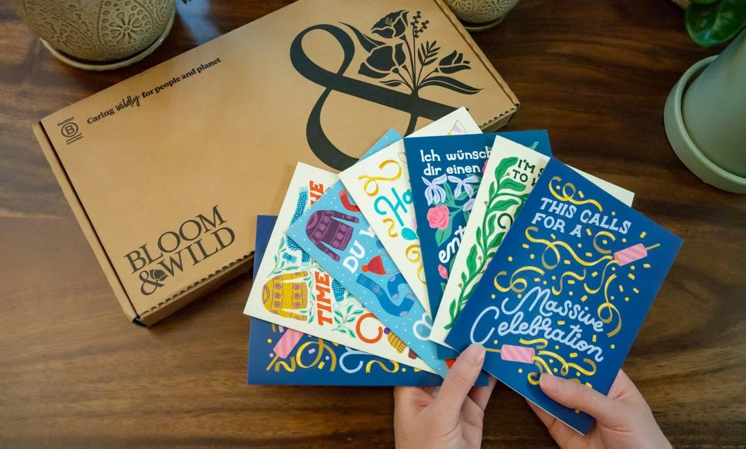 A person holding a set of colorful greeting cards in front of a brown cardboard box labeled 'Bloom & Wild' on a wooden table. The cards have various celebratory messages, and the box features a black floral logo.