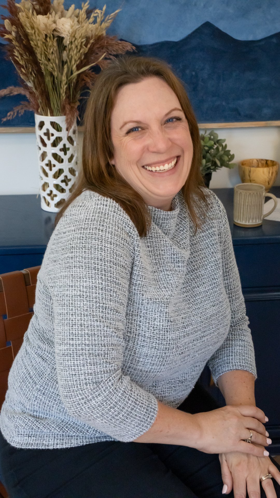 A woman with brown hair smiling, wearing a light patterned sweater, sitting at a table with a background of a blue painting and decorative items like a vase with dried plants, a small potted plant, a mug, and a wooden bowl.