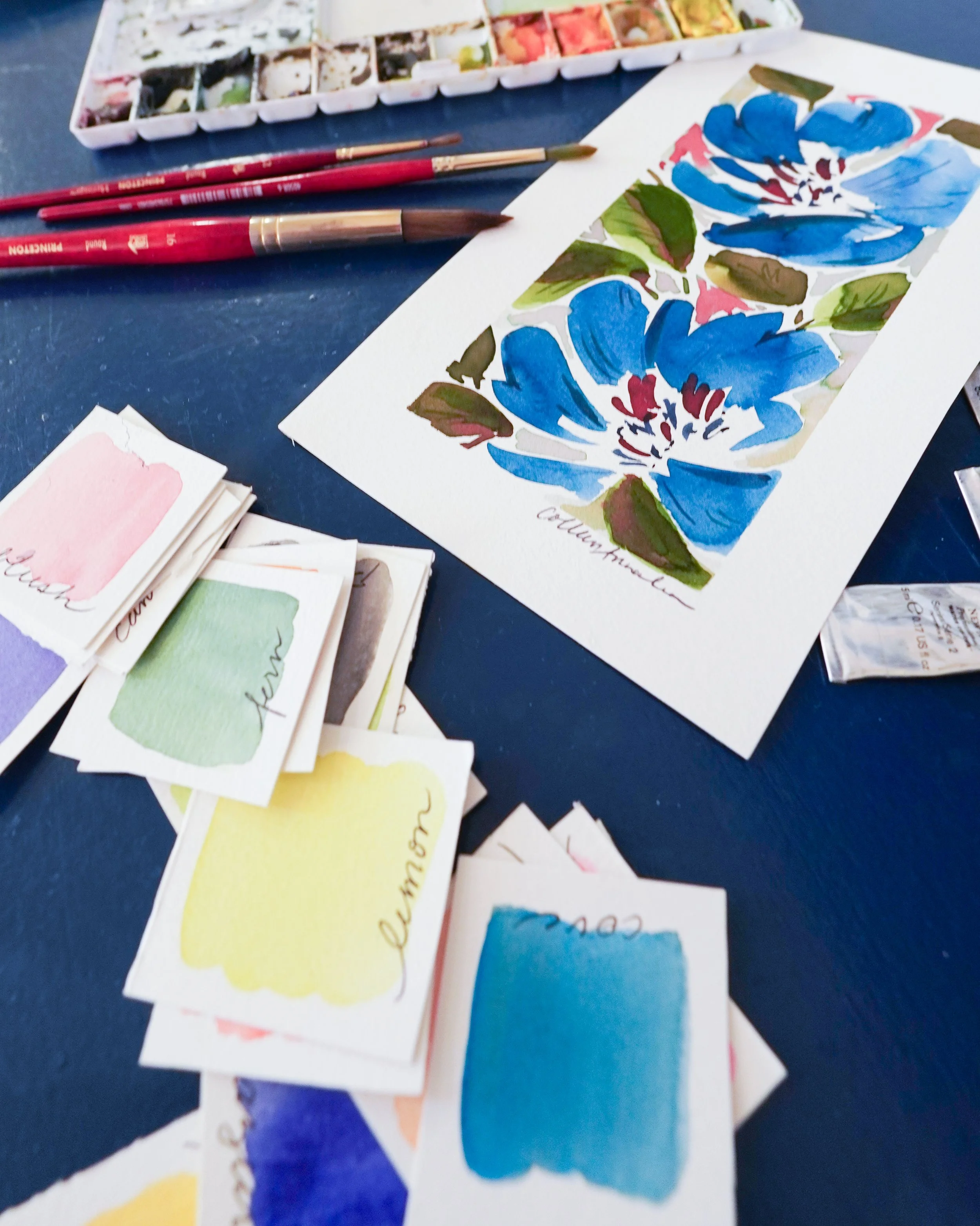 Watercolor painting of blue flowers with green leaves on white paper, watercolor palette with various color paints, and paintbrushes placed on a dark blue surface.