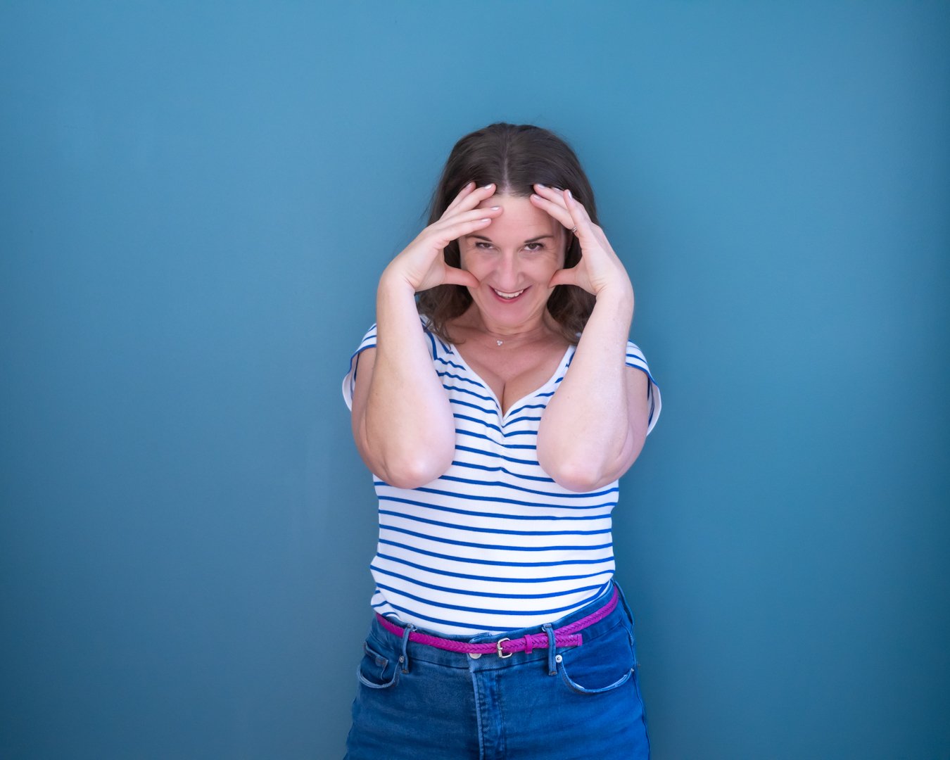 Smiling woman standing against a blue background, wearing a striped t-shirt and blue jeans, holding her head with both hands.