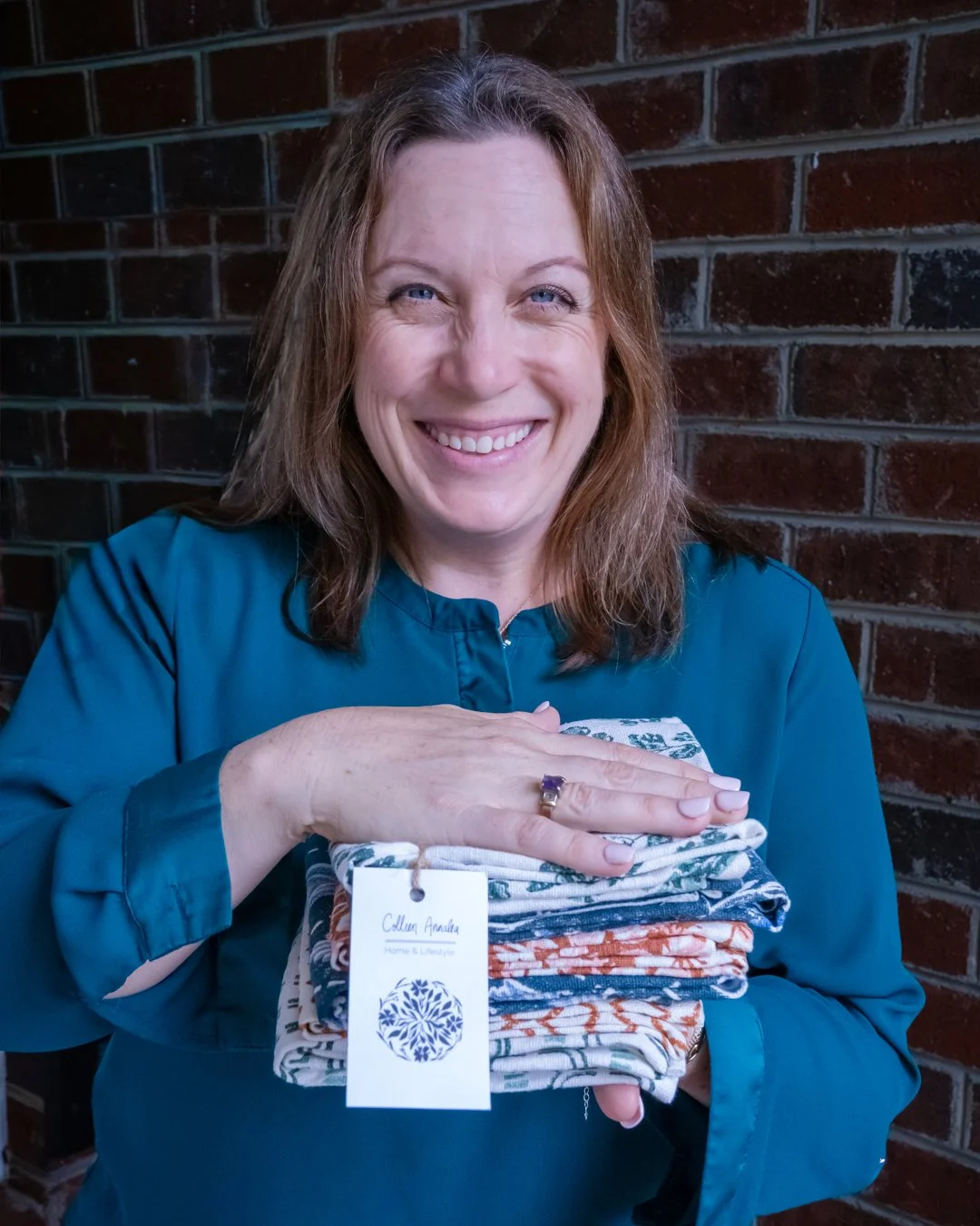 A woman with shoulder-length brown hair smiling and holding a stack of folded clothes with a tag that reads 'Callen Angela, Home & Lifestyle', standing in front of a brick wall.