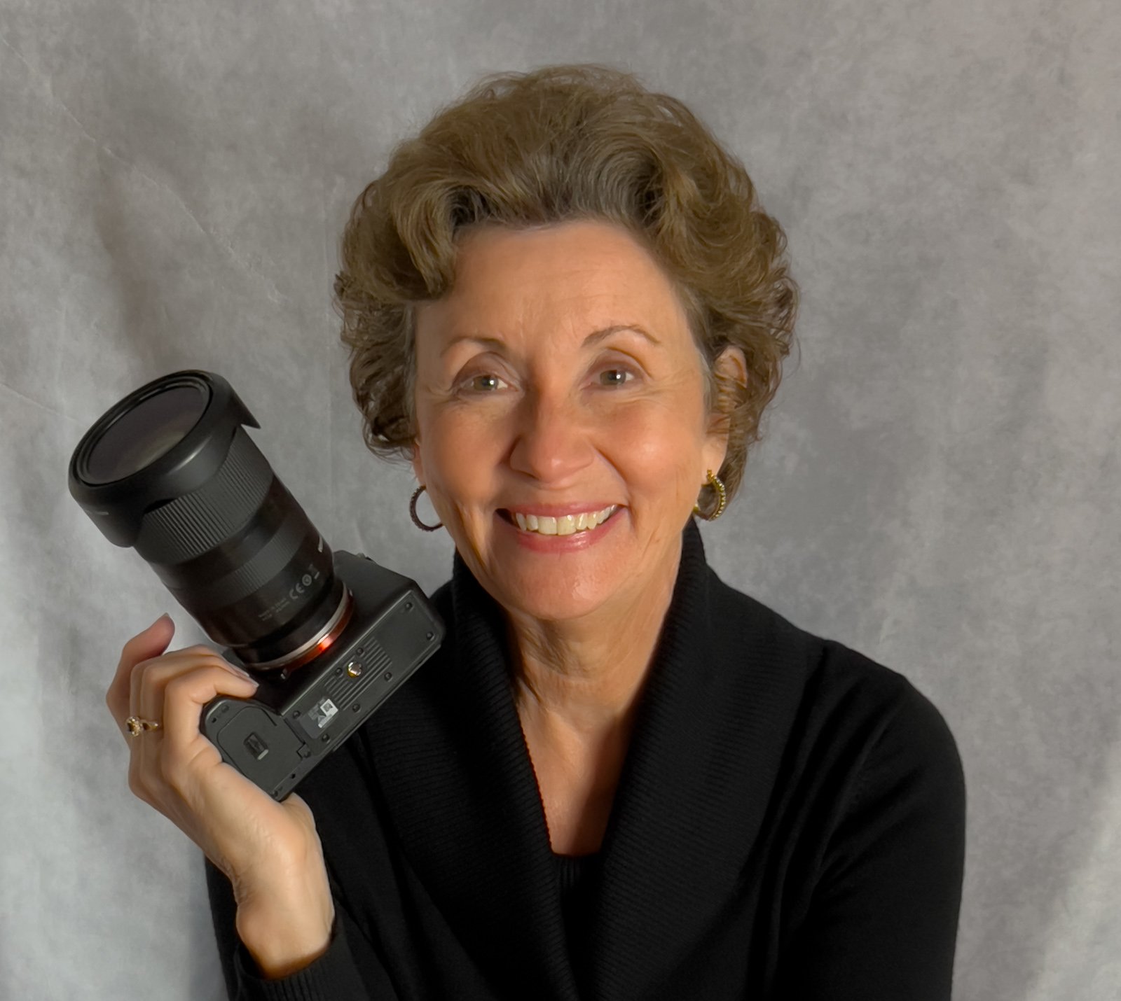 Portrait of a happy female photographer with short, curly hair, smiling, holding a camera with a large lens. She wears gold earrings and a black top, sitting in front of a gray, textured background.