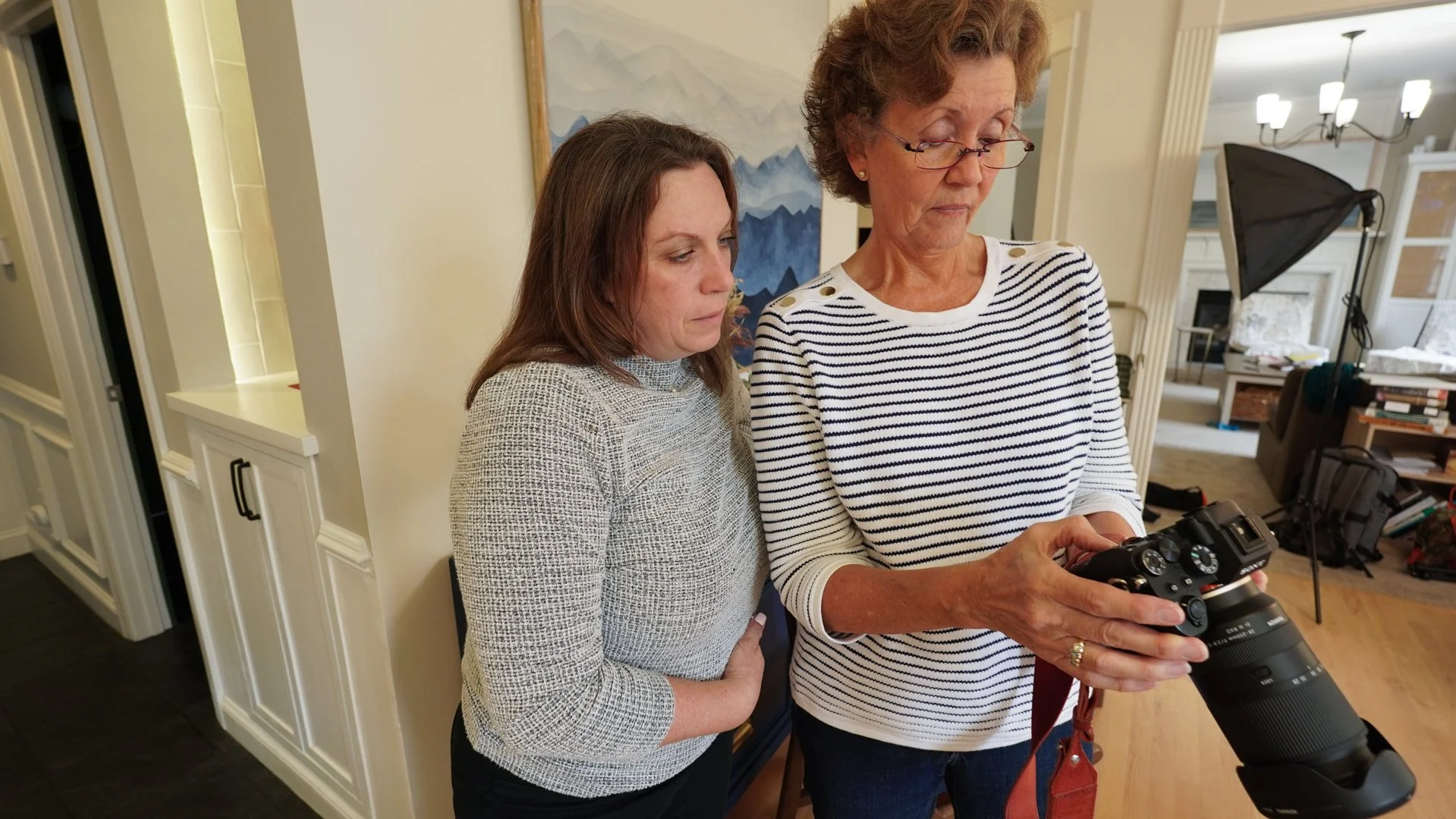 Two women are standing together indoors, looking at a camera that one of them is holding. The woman on the right has short, curly hair, wears glasses, and a striped shirt. The woman on the left has shoulder-length hair and a patterned sweater. The background features a living room with books, a shelving unit, a chandelier, and a painting of mountains.