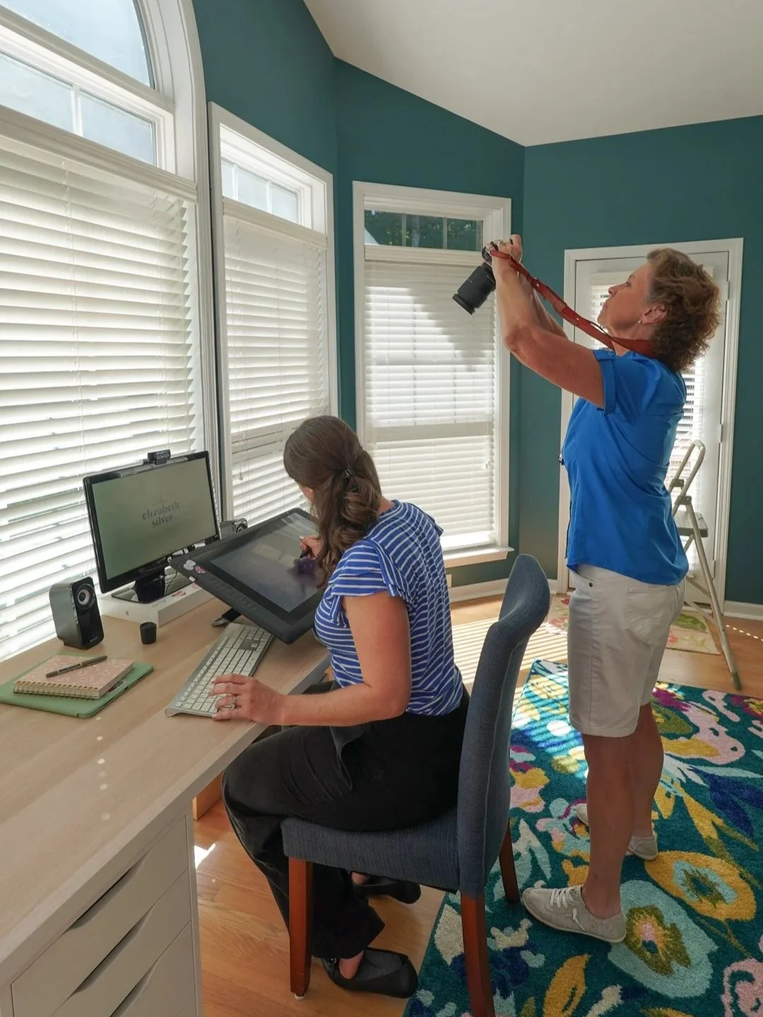 Two women in a well-lit room with large windows, one sitting at a desk working on a graphics tablet, and the other standing behind her taking photographs with a camera.