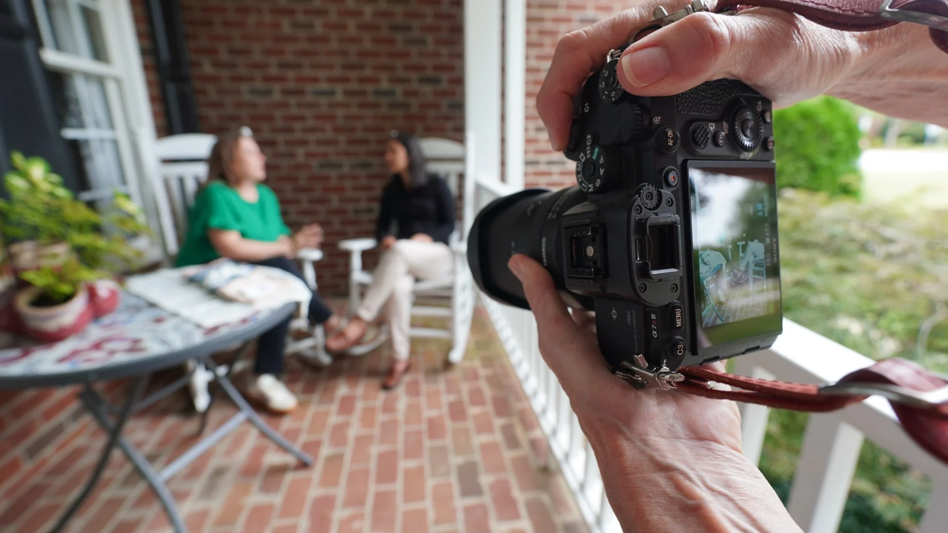 Photographer capturing two women sitting and talking on a porch with brick walls, plants, and outdoor furniture.