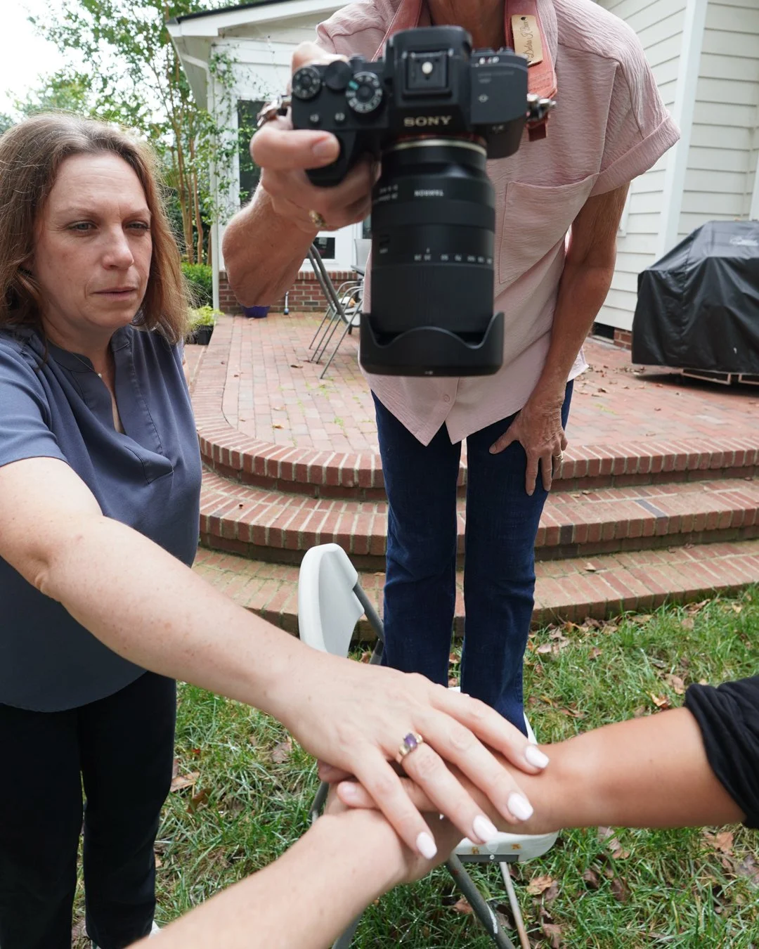 Two women are participating in a ceremonial hand blessing outside a house. One woman is sitting, and the other is standing with a camera aimed at them. They are holding hands with their palms together. The setting includes a brick patio, a house in the background, and some outdoor furniture.