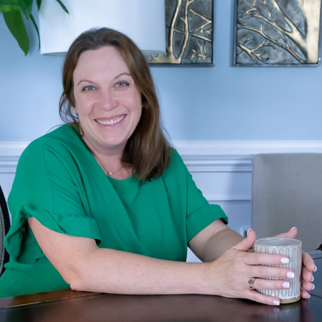 Woman with brown hair, smiling, wearing a green top, sitting at a table with a coffee mug in her hands, in a room with framed abstract art and a beige chair.