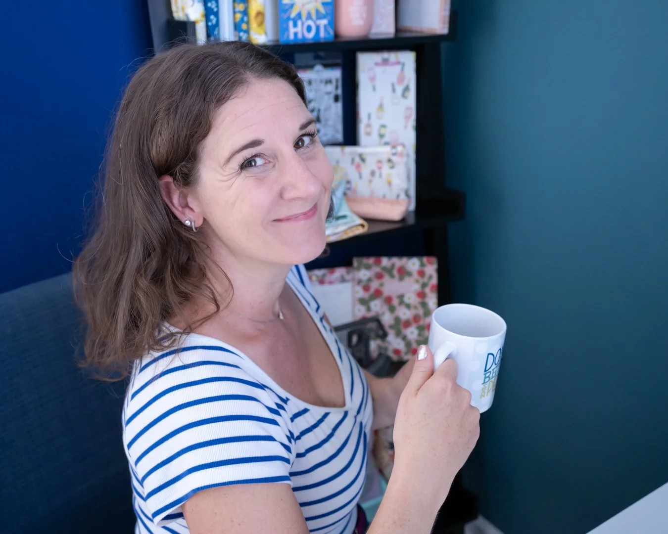 A woman with shoulder-length brown hair, wearing a white and blue striped shirt, sitting in an office, holding a white mug with a design, smiling at the camera. There are shelves with colorful boxes and items in the background.