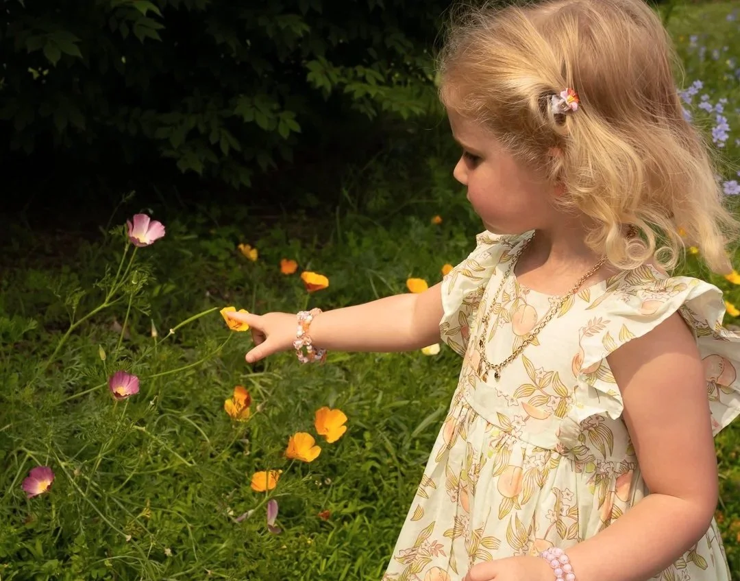 A young girl with curly blonde hair, wearing a floral dress and jewelry, touching purple and orange flowers in a garden.