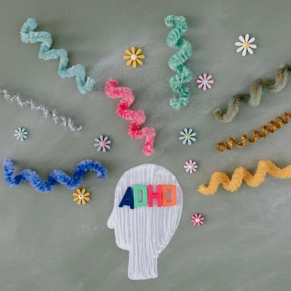 Decorative paper cutout of a human head silhouette with colorful foam letters spelling 'ADHD' inside it. Surrounding the head are colorful pipe cleaners, small foam flower decorations, and tinsel on a plain background.