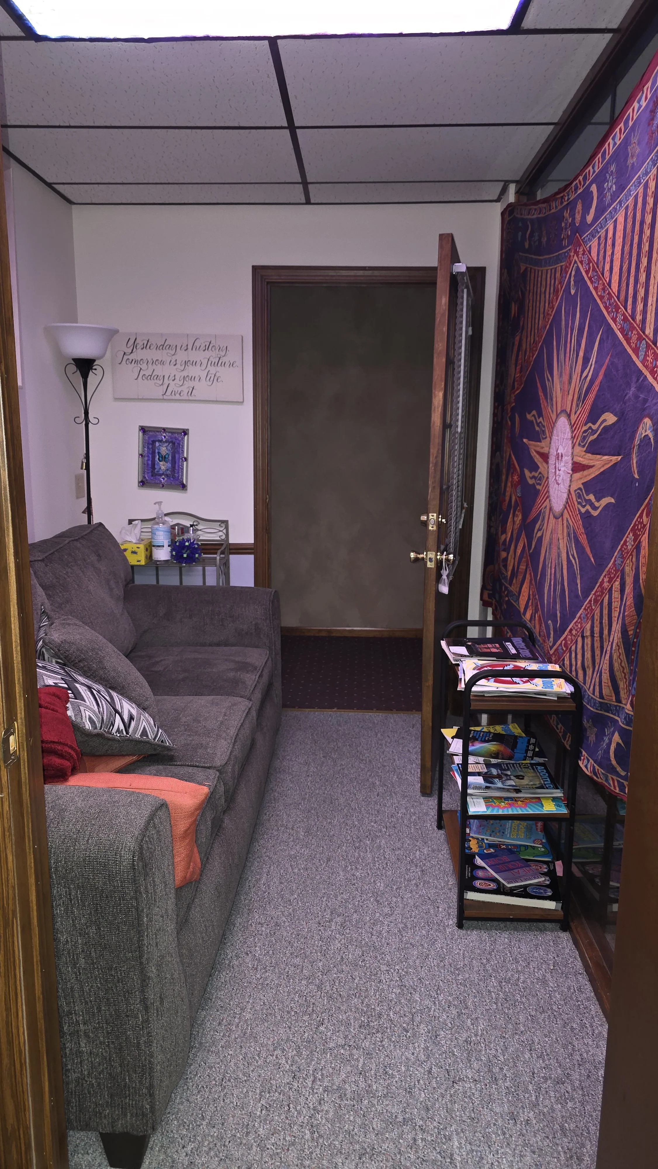 A waiting room with an open door, furnished with a comfy grey couch, a stand lamp, shelves with magazines and a wall tapestry with a sun design.