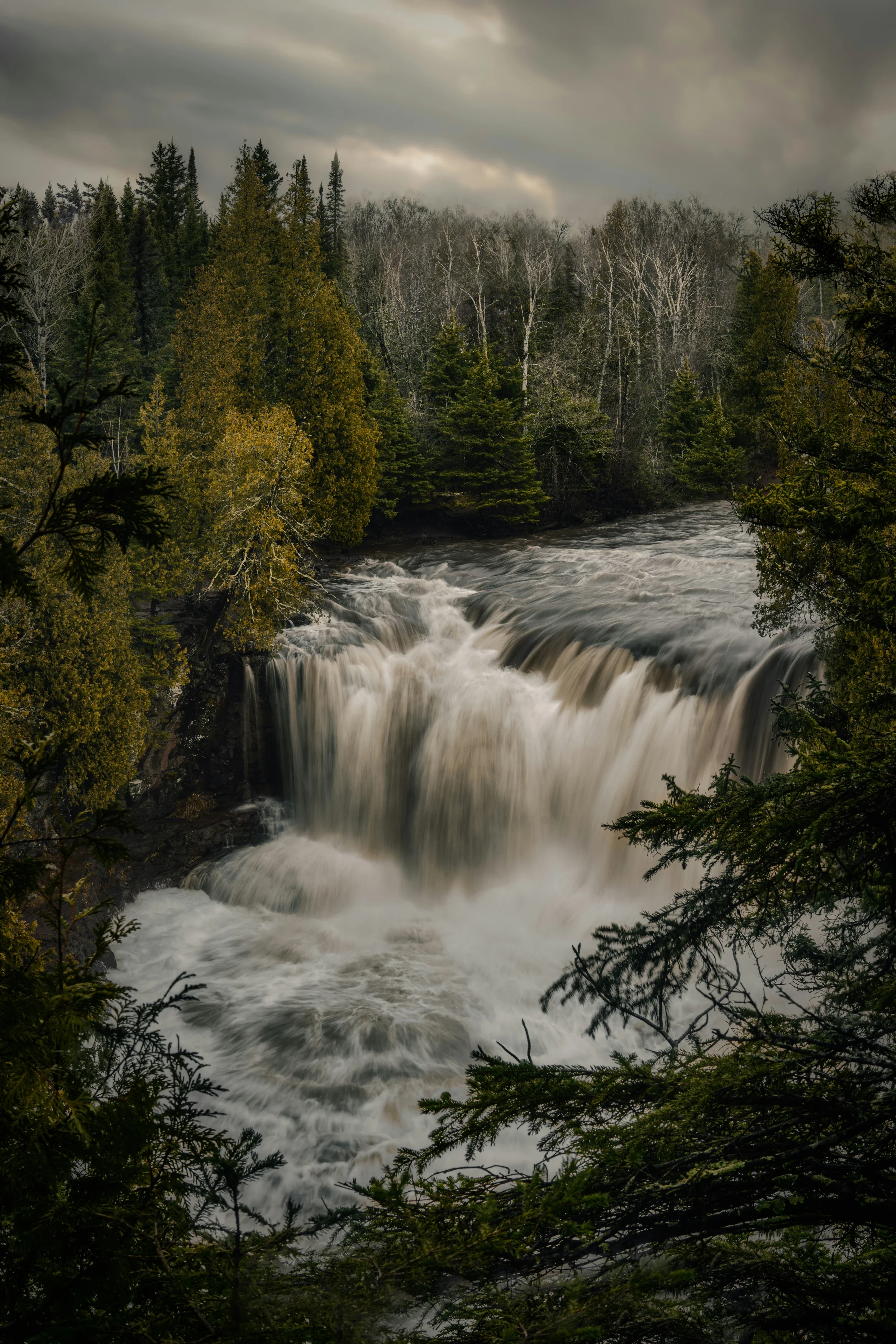 A waterfall flowing through a forest of evergreen and deciduous trees on a cloudy day.