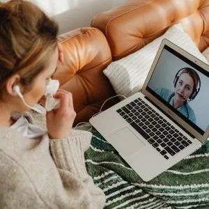 A woman participating in a video call on her laptop, sitting on a bed with a pillow and blanket.