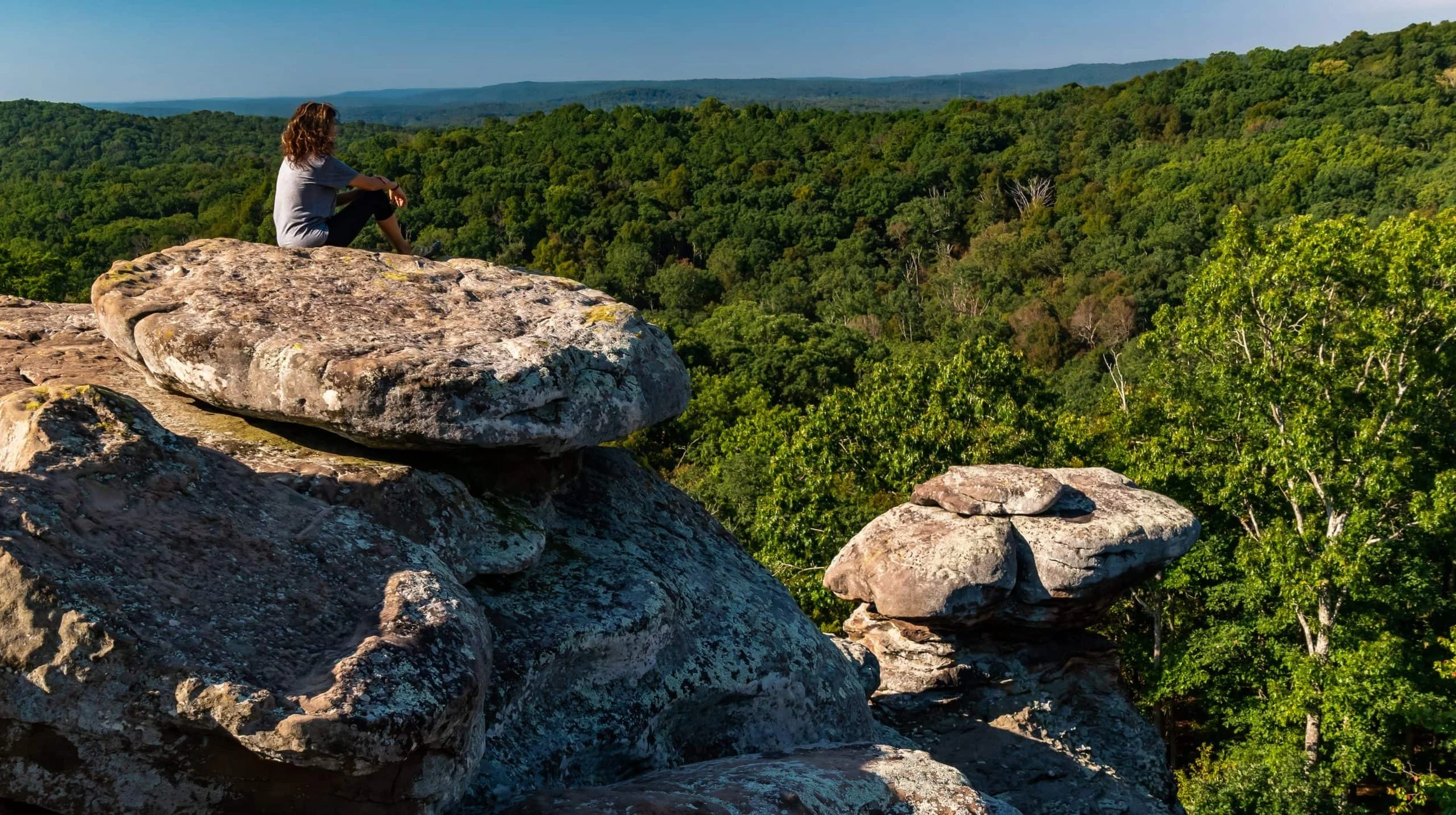 A woman sitting on rocks overlooking a vast green forest with distant hills.