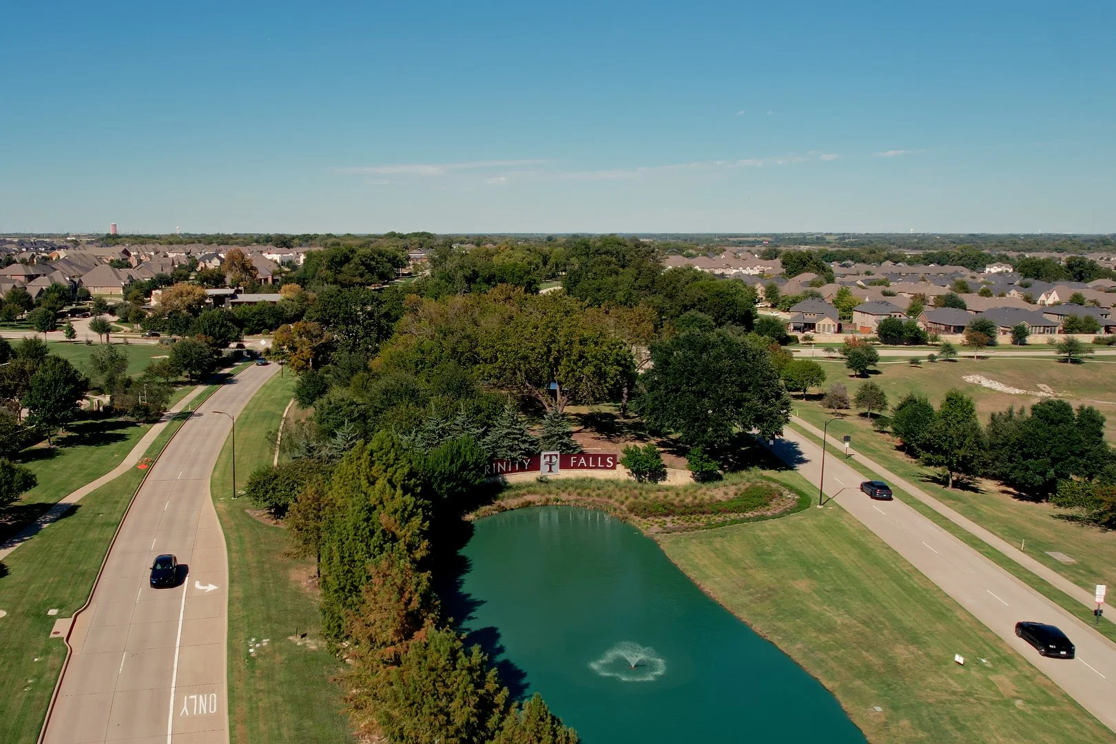 Drone image of the entrance of Trinity Falls in McKinney, Texas featuring landscaped waterfalls, stone bridges, walking paths, community lake views, and upscale master-planned neighborhood homes.