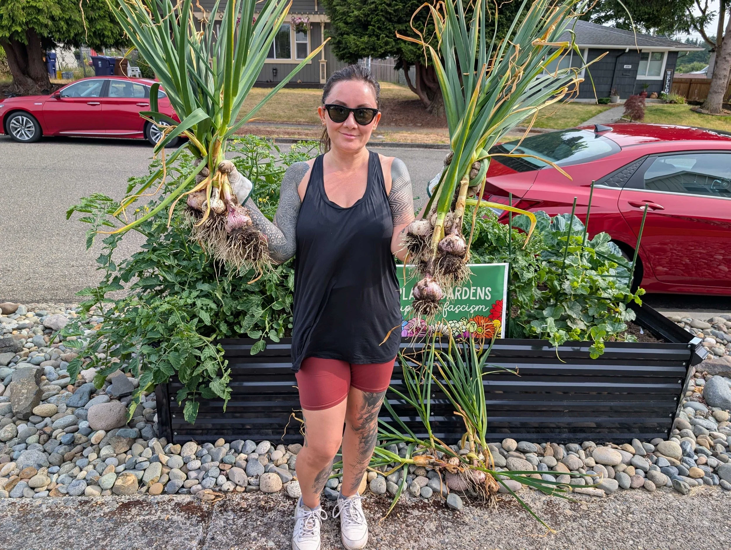 An image of Linda in front of one of her garden beds holding up her garlic harvest