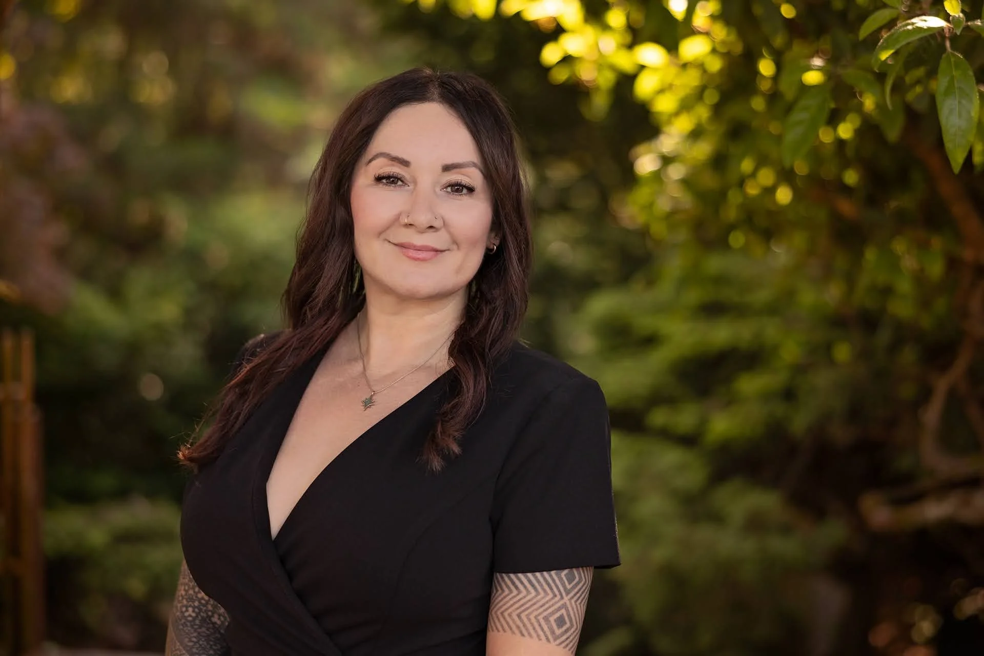 A woman with long brown hair, tattoos on her arms, wearing a black dress, standing outdoors with a background of green trees and sunlight filtering through leaves.