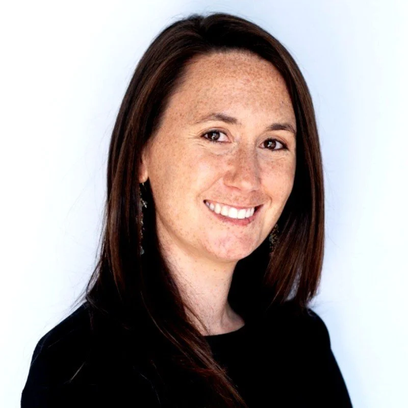 A woman with straight auburn hair, fair skin with freckles, smiling and looking at the camera, wearing earrings and a black top against a plain white background.