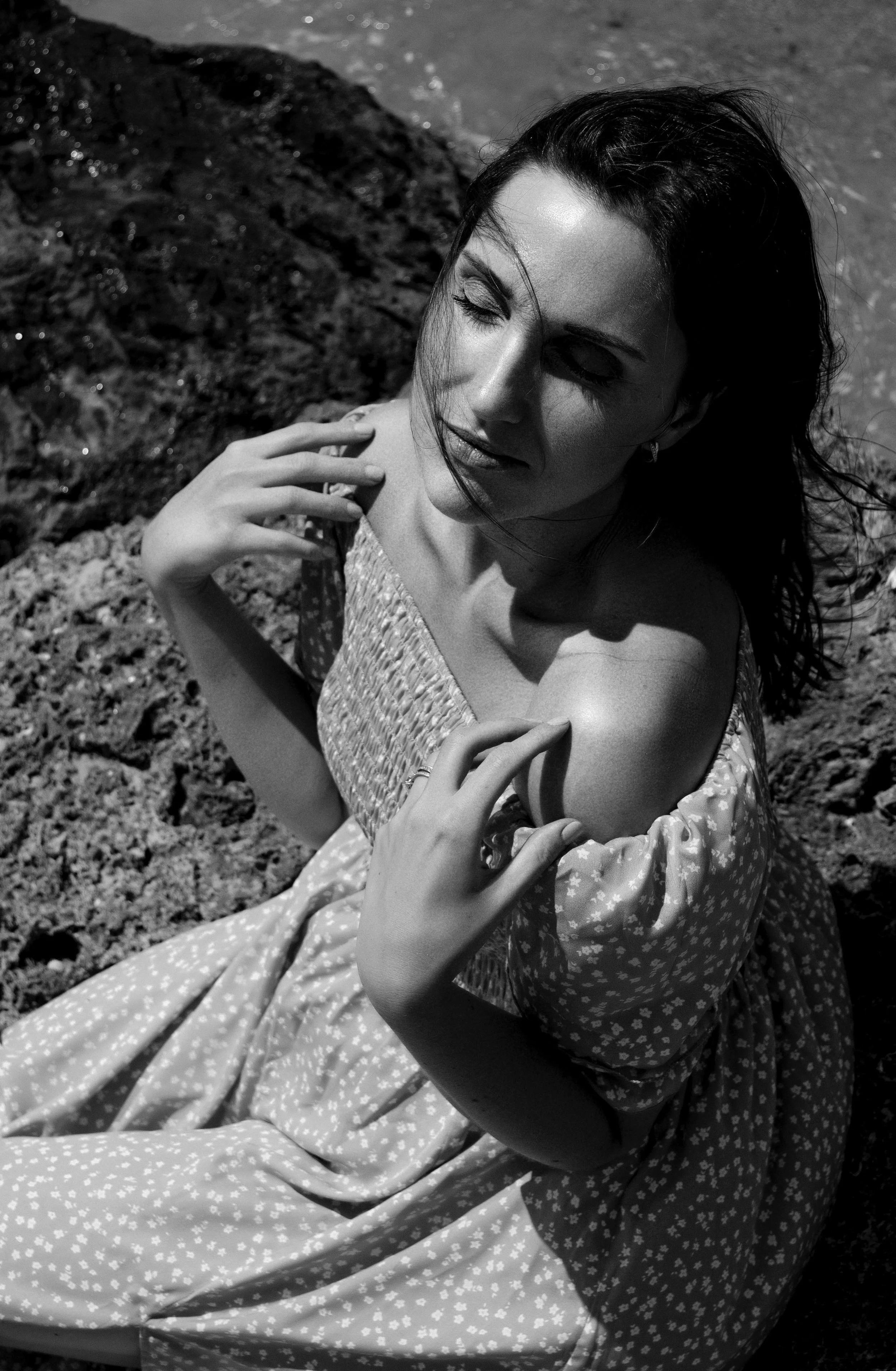 Black and white photo of a woman with long dark hair, sitting outdoors on rocks near water, with her eyes closed and hands gently resting on her shoulder.