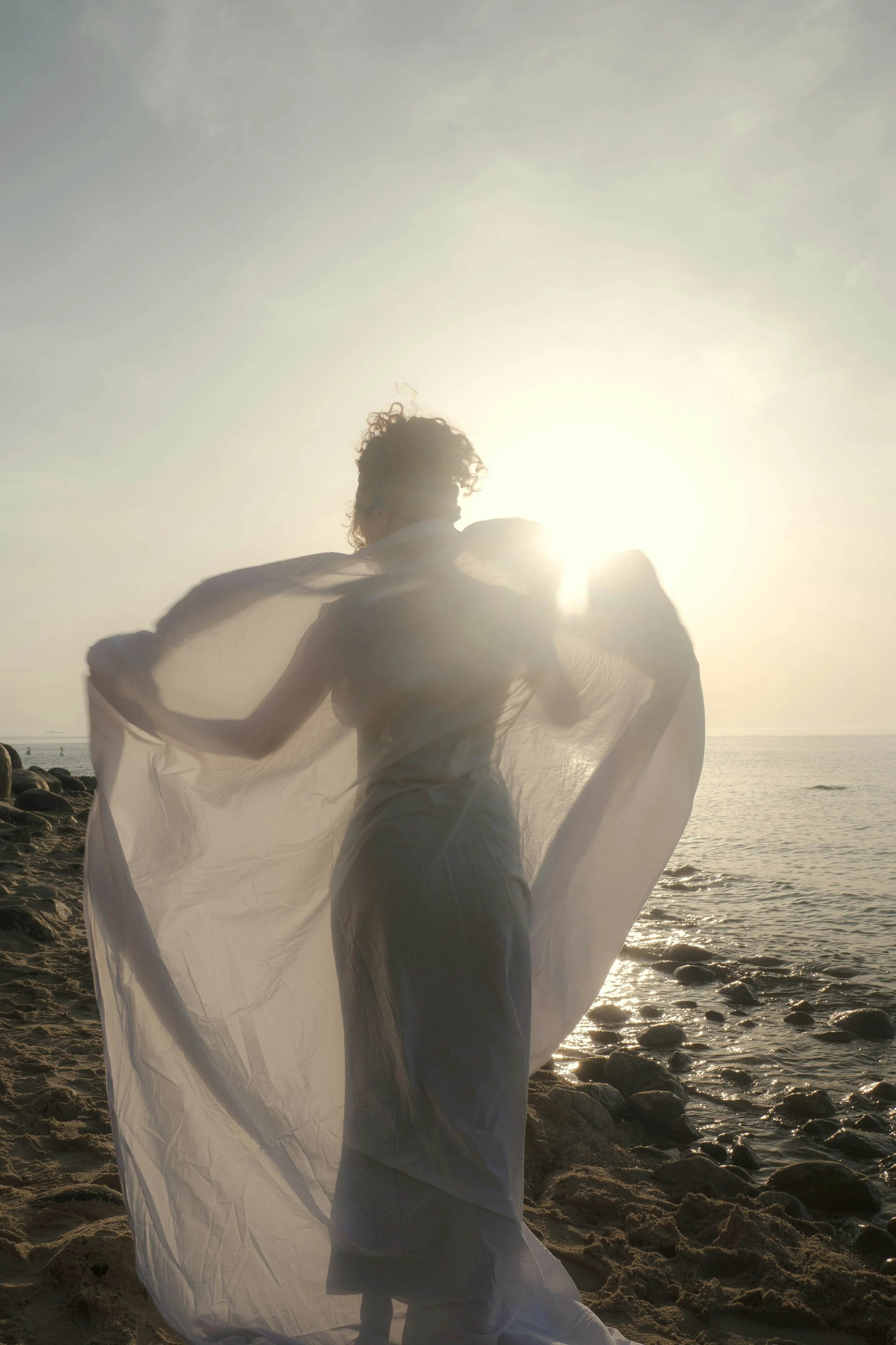 A person with curly hair dressed in flowing white fabric stands on a rocky beach at sunset, facing the ocean with the sun shining brightly behind them.