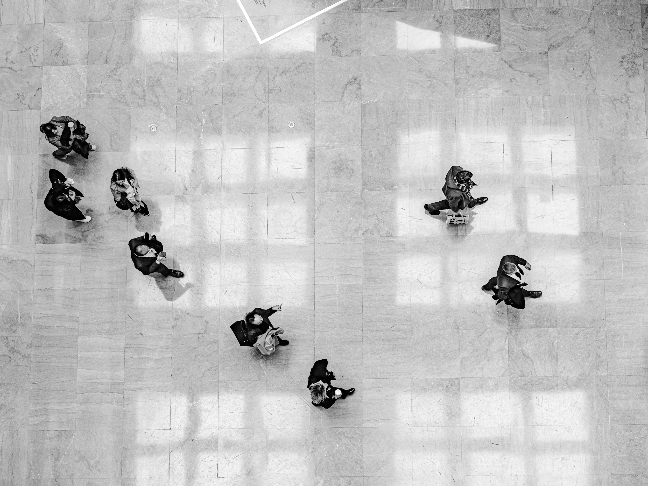 An overhead black-and-white photograph of people walking in a large, tiled indoor space. The individuals are scattered, some walking alone, others in pairs or small groups, with shadows cast on the floor.