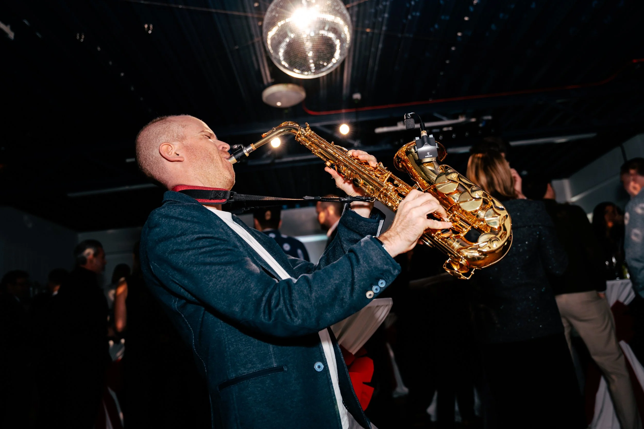 A man playing the saxophone at an indoor event or party, surrounded by other people in the background.