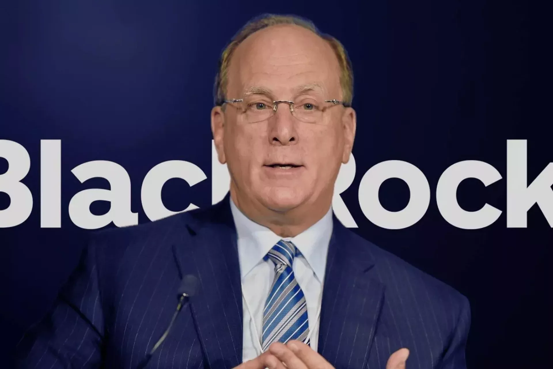 An older man with glasses and light brown hair, wearing a dark navy suit, white shirt, and a striped blue and white tie, speaking at a podium with a dark background featuring large white text that reads 'Blackrock'.