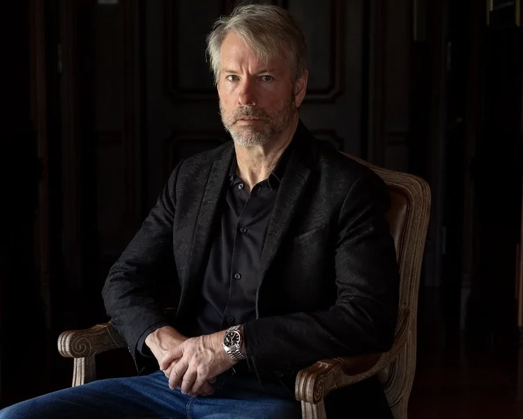 A middle-aged man with gray hair and beard, wearing a black shirt and blazer, sitting on a wooden chair in a dimly lit room with dark wood paneling background.