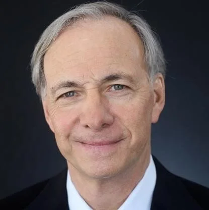 Headshot of a middle-aged man with gray hair and blue eyes, wearing a dark suit and white shirt, smiling at the camera against a black background.
