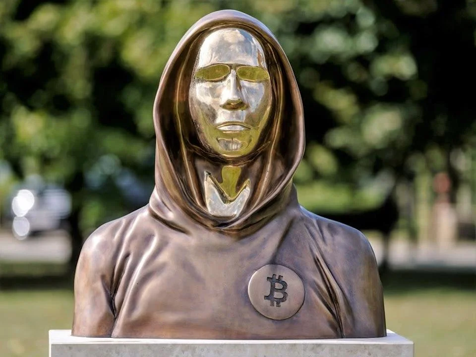 Bronze sculpture of a person wearing a hood with a gold-colored mask resembling a human face, with a Bitcoin symbol on their chest, outdoors with blurry green trees in the background.
