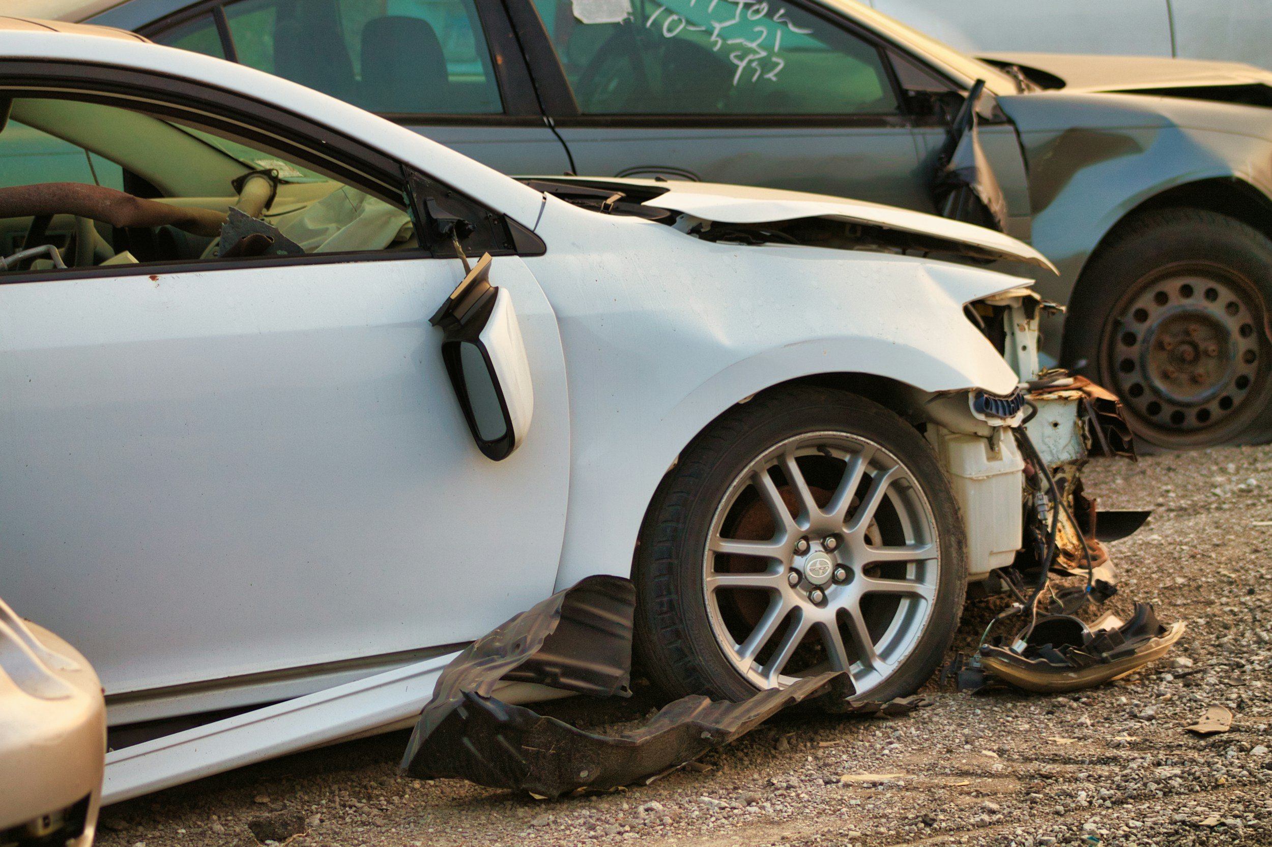 Two damaged cars, one white and one gray, parked outdoors on a dirt surface; the white car has significant front-end damage with parts hanging and exposed.