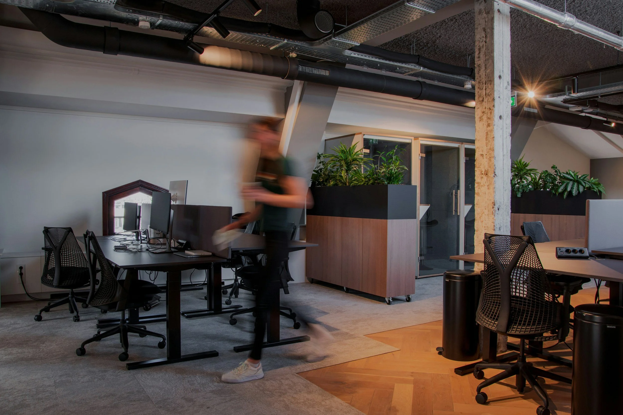 Modern coworking space with tables, chairs, computer monitors, indoor plants, and an exposed ceiling with black ductwork and industrial lighting.