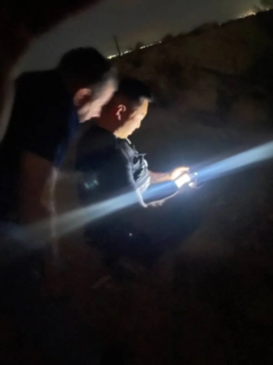 Two young men standing outdoors at night, one of them looking at a phone with a bright flashlight or screen illuminating his face, with a dark background and distant lights visible.