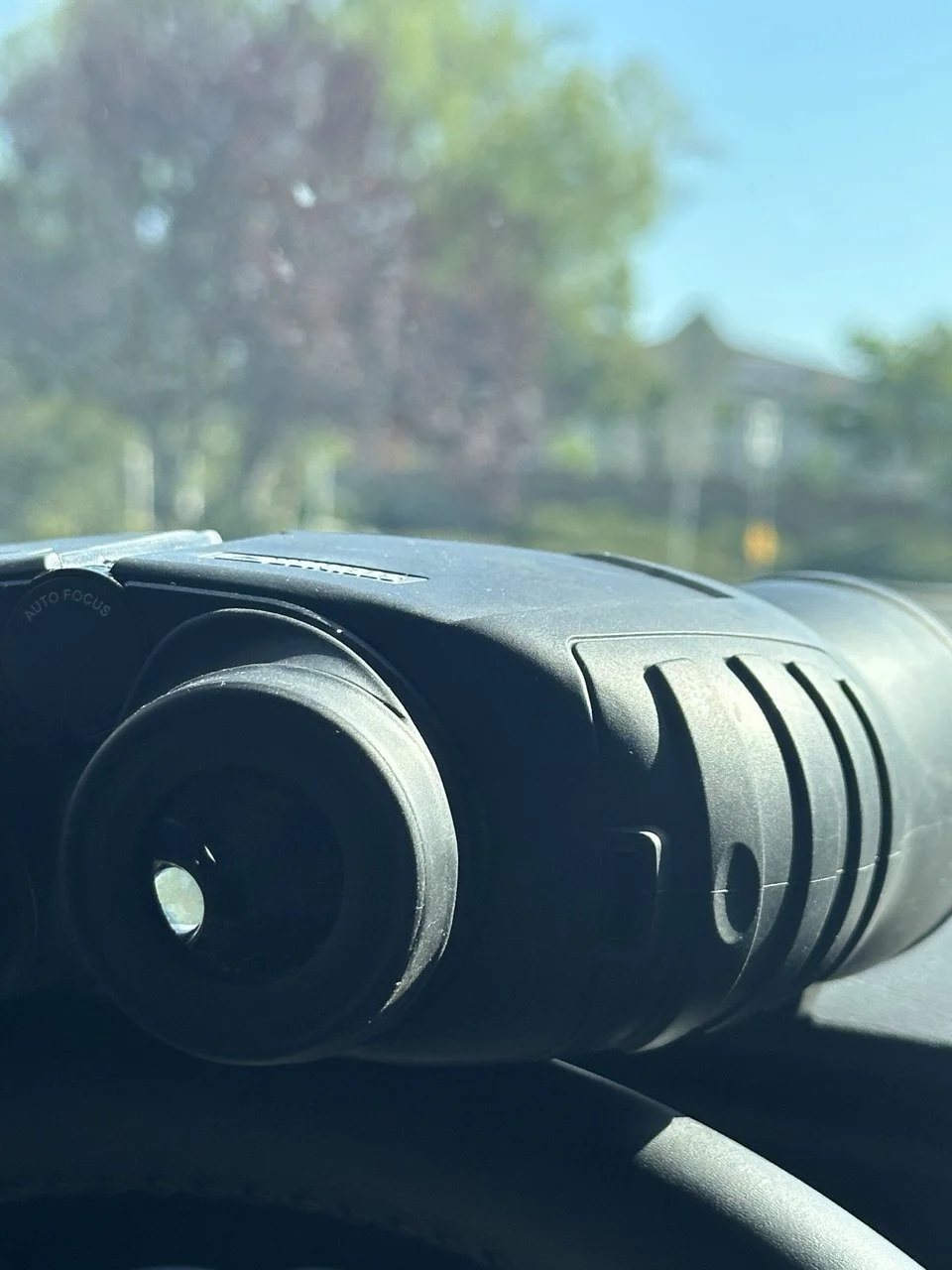 Black binoculars on a car dashboard with a blurry view of trees and a house outside the windshield.