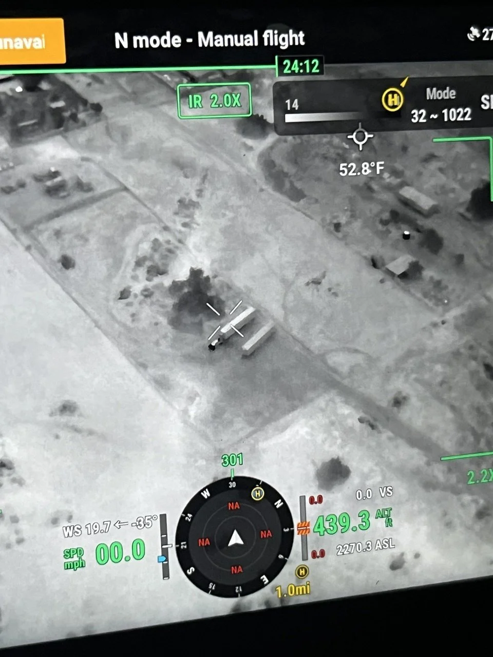 A drone's infrared camera view showing a ground scene with some buildings, vehicles, and a person lying on the ground near a structure, with flight data overlay including altitude, speed, temperature, and navigation information.