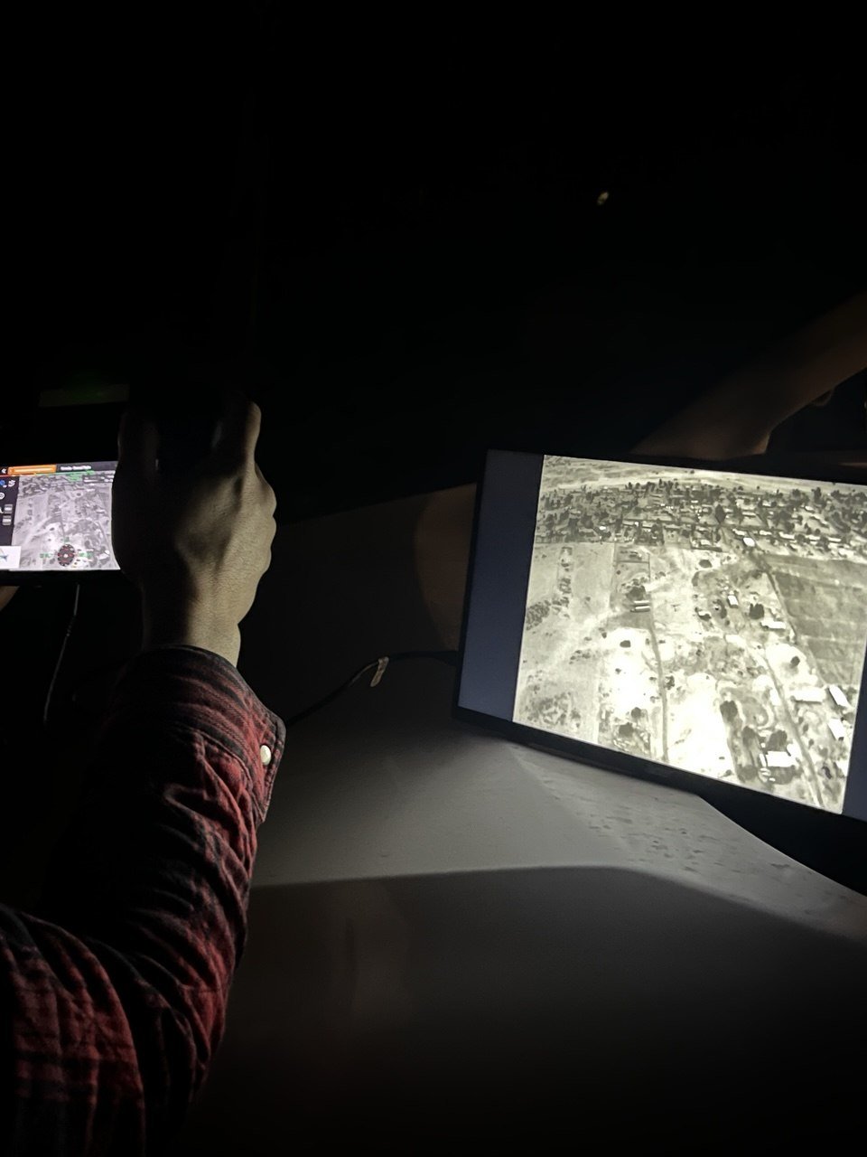 A person holding a phone in front of a car dashboard at night, with a tablet on the dashboard displaying a satellite view of a landscape with buildings and roads.