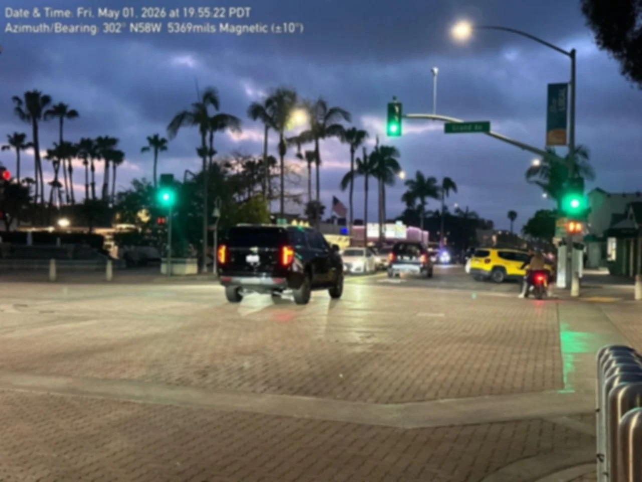 A busy intersection in a city during dusk with palm trees, traffic lights, and several cars waiting at the red light. The sky is cloudy and the streetlights are on.