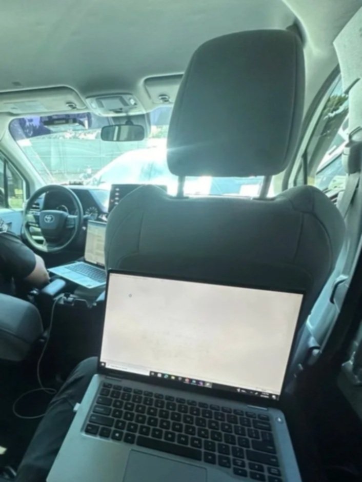 Inside a vehicle, two laptops on seats, one person working to the left and the driver on the front left, with other cars visible outside through the windows.