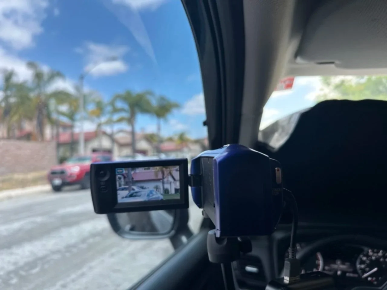 View of a car dashboard with a dash camera mounted on the windshield, capturing the street scene outside with a blurred red car, trees, and houses under a blue sky.