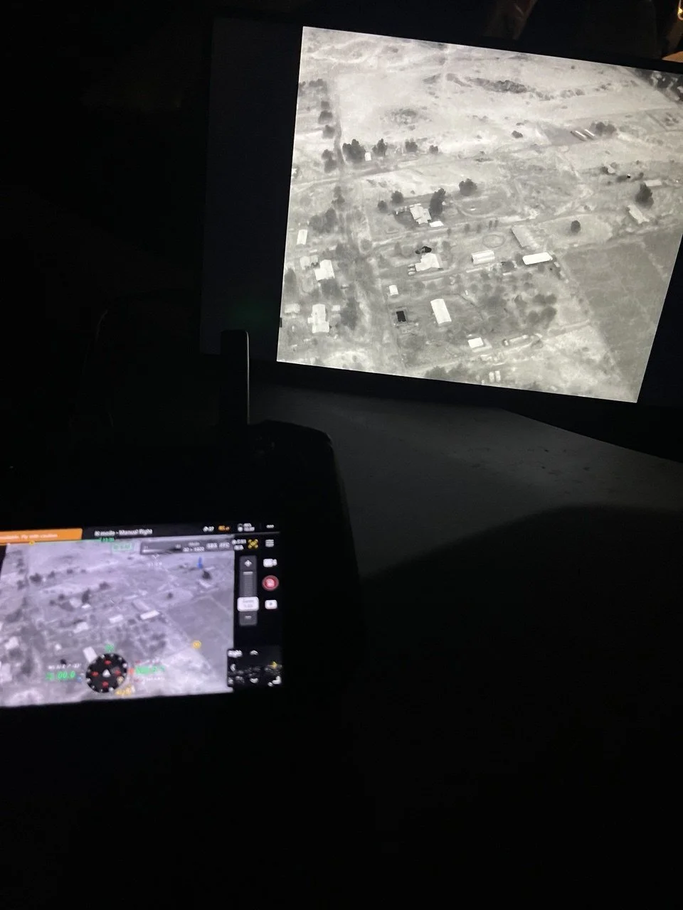 Aerial view of a rural area with scattered buildings and trees, seen on a screen in a dark environment.