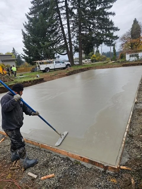 Construction worker smoothing freshly poured concrete on a flat surface with a long-handled tool, with trees and trucks in the background.