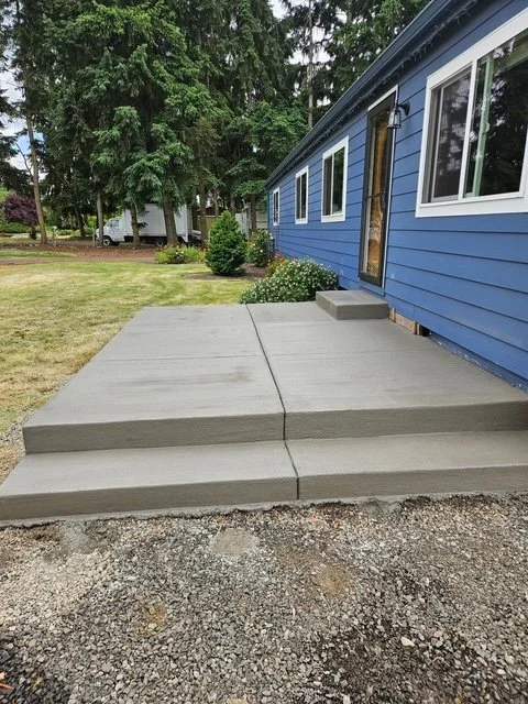 A newly poured concrete porch with steps in front of a blue house with windowed siding, surrounded by a lawn and trees.