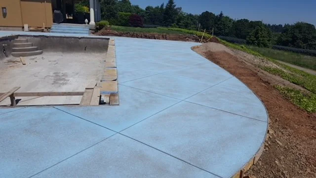 Concrete pool deck under construction with a curved shape, surrounded by soil and grass, and a pool foundation in the early stages of building.